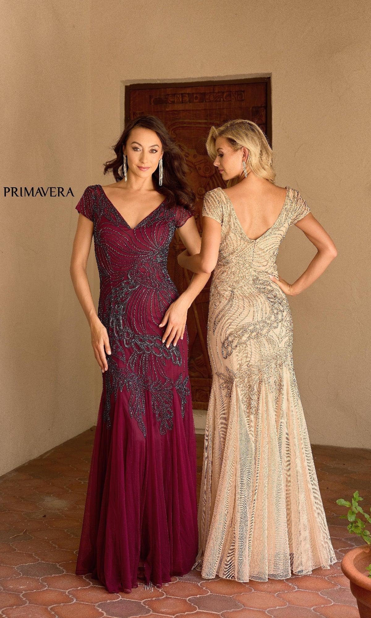 Two women model the Primavera 13148 long formal dress: one faces forward in a burgundy v-neck gown, the other faces backward in a champagne beaded version. Both stand on a tiled floor.